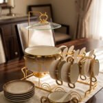 Luxury gradient brown ceramic soup tureen pot with glass lid, gold warmer stand, hanging double-handled soup bowls, saucers, and white spoons set on a wooden dining table.
