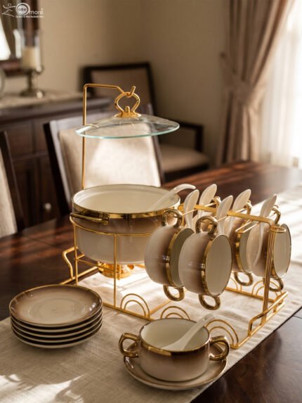 Luxury gradient brown ceramic soup tureen pot with glass lid, gold warmer stand, hanging double-handled soup bowls, saucers, and white spoons set on a wooden dining table.