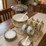 Luxury white ceramic soup tureen set with gold accents, hanging glass lid, matching soup bowls, saucers, and spoons on a gold metal stand.