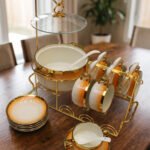 Ceramic soup tureen set with orange gradient rims and gold accents, including hanging soup cups, saucers, and spoons organized on a gold metal rack.