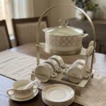 Luxury white and gold geometric pattern ceramic soup tureen set with matching mugs, spoons, and silver metallic rack on a marble kitchen counter