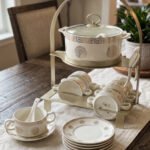 White ceramic soup tureen with glass lid, matching dual-handled soup bowls, saucers, and spoons organized on a two-tier gold metal rack.