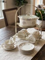 White ceramic soup tureen with glass lid, matching dual-handled soup bowls, saucers, and spoons organized on a two-tier gold metal rack.
