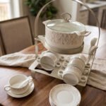 Ceramic soup tureen with clear glass lid, matching soup bowls with handles, saucers, and spoons arranged on a two-tier metal stand on a wooden dining table.