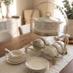 Luxury ceramic soup tureen set featuring a patterned soup pot, soup bowls with handles, matching saucers, spoons, and a gold metal stand on a wooden dining table.
