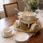 Luxury white and gold ceramic soup tureen set with warming stand, matching bowls, saucers, spoons, and glass lid on a wooden table.