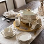Ceramic soup tureen set with glass lid, matching double-handled bowls, saucers, spoons, and a gold metal warming stand on a dining table setting.