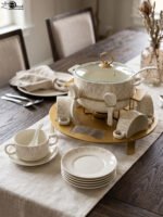 Ceramic soup tureen set with glass lid, matching double-handled bowls, saucers, spoons, and a gold metal warming stand on a dining table setting.