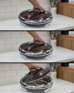 An elegant oval-shaped insulated hotpot in dark brown with a silver stainless steel interior and decorative studded handles, open with the lid resting on a polished wooden dining table.