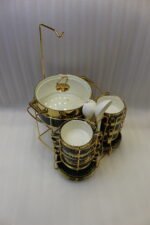 Luxury emerald green ceramic soup tureen set with gold accents, glass lid, warmer stand, stacked double-handled cups, saucers, and white spoons on a marble countertop.