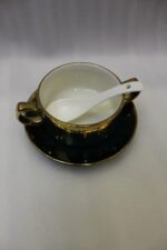 Luxury emerald green ceramic soup tureen set with gold accents, glass lid, warmer stand, stacked double-handled cups, saucers, and white spoons on a marble countertop.