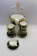 Luxury emerald green ceramic soup tureen set with gold accents, glass lid, warmer stand, stacked double-handled cups, saucers, and white spoons on a marble countertop.