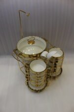 White ceramic soup tureen and stacked double-handled soup bowls with gold rim accents on a gold wire stand on a dining table.