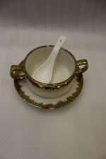 White ceramic soup tureen and stacked double-handled soup bowls with gold rim accents on a gold wire stand on a dining table.