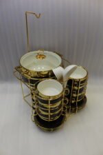 Luxury black and gold ceramic soup set with double-handled cups, covered soup tureen, white spoons, and gold metal display rack on a marble kitchen counter.