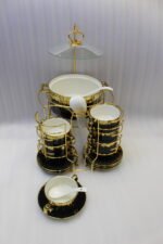 Luxury black and gold ceramic soup set with double-handled cups, covered soup tureen, white spoons, and gold metal display rack on a marble kitchen counter.
