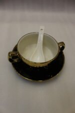 Luxury black and gold ceramic soup set with double-handled cups, covered soup tureen, white spoons, and gold metal display rack on a marble kitchen counter.