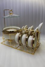 Luxury gradient brown ceramic soup tureen pot with glass lid, gold warmer stand, hanging double-handled soup bowls, saucers, and white spoons set on a wooden dining table.