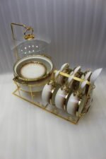 Luxury gradient brown ceramic soup tureen pot with glass lid, gold warmer stand, hanging double-handled soup bowls, saucers, and white spoons set on a wooden dining table.