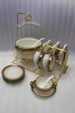 Luxury gradient brown ceramic soup tureen pot with glass lid, gold warmer stand, hanging double-handled soup bowls, saucers, and white spoons set on a wooden dining table.