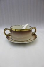 Luxury gradient brown ceramic soup tureen pot with glass lid, gold warmer stand, hanging double-handled soup bowls, saucers, and white spoons set on a wooden dining table.