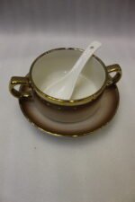 Luxury gradient brown ceramic soup tureen pot with glass lid, gold warmer stand, hanging double-handled soup bowls, saucers, and white spoons set on a wooden dining table.
