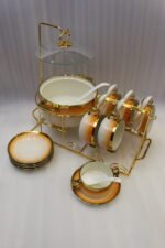 Ceramic soup tureen set with orange gradient rims and gold accents, including hanging soup cups, saucers, and spoons organized on a gold metal rack.