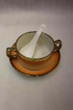 Ceramic soup tureen set with orange gradient rims and gold accents, including hanging soup cups, saucers, and spoons organized on a gold metal rack.