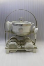 White ceramic soup tureen with glass lid, matching dual-handled soup bowls, saucers, and spoons organized on a two-tier gold metal rack.