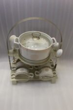 White ceramic soup tureen with glass lid, matching dual-handled soup bowls, saucers, and spoons organized on a two-tier gold metal rack.