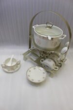 White ceramic soup tureen with glass lid, matching dual-handled soup bowls, saucers, and spoons organized on a two-tier gold metal rack.