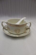 White ceramic soup tureen with glass lid, matching dual-handled soup bowls, saucers, and spoons organized on a two-tier gold metal rack.