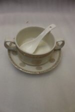 White ceramic soup tureen with glass lid, matching dual-handled soup bowls, saucers, and spoons organized on a two-tier gold metal rack.