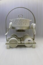 Ceramic soup tureen with clear glass lid, matching soup bowls with handles, saucers, and spoons arranged on a two-tier metal stand on a wooden dining table.