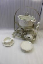 Ceramic soup tureen with clear glass lid, matching soup bowls with handles, saucers, and spoons arranged on a two-tier metal stand on a wooden dining table.