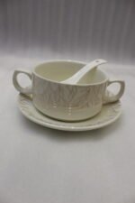 Ceramic soup tureen with clear glass lid, matching soup bowls with handles, saucers, and spoons arranged on a two-tier metal stand on a wooden dining table.