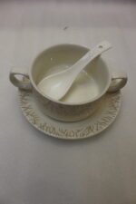 Ceramic soup tureen with clear glass lid, matching soup bowls with handles, saucers, and spoons arranged on a two-tier metal stand on a wooden dining table.