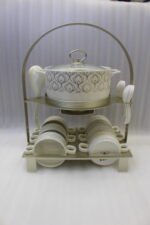 Luxury ceramic soup tureen set featuring a patterned soup pot, soup bowls with handles, matching saucers, spoons, and a gold metal stand on a wooden dining table.