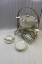 Luxury ceramic soup tureen set featuring a patterned soup pot, soup bowls with handles, matching saucers, spoons, and a gold metal stand on a wooden dining table.