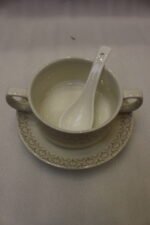 Luxury ceramic soup tureen set featuring a patterned soup pot, soup bowls with handles, matching saucers, spoons, and a gold metal stand on a wooden dining table.
