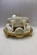 Luxury white and gold ceramic soup tureen set with warming stand, matching bowls, saucers, spoons, and glass lid on a wooden table.