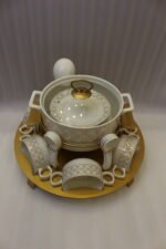 Luxury white and gold ceramic soup tureen set with warming stand, matching bowls, saucers, spoons, and glass lid on a wooden table.