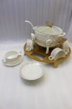 Luxury white and gold ceramic soup tureen set with warming stand, matching bowls, saucers, spoons, and glass lid on a wooden table.