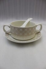 Luxury white and gold ceramic soup tureen set with warming stand, matching bowls, saucers, spoons, and glass lid on a wooden table.