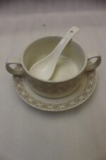 Luxury white and gold ceramic soup tureen set with warming stand, matching bowls, saucers, spoons, and glass lid on a wooden table.