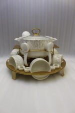 Ceramic soup tureen set with glass lid, matching double-handled bowls, saucers, spoons, and a gold metal warming stand on a dining table setting.
