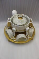 Ceramic soup tureen set with glass lid, matching double-handled bowls, saucers, spoons, and a gold metal warming stand on a dining table setting.