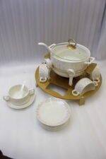 Ceramic soup tureen set with glass lid, matching double-handled bowls, saucers, spoons, and a gold metal warming stand on a dining table setting.