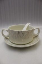Ceramic soup tureen set with glass lid, matching double-handled bowls, saucers, spoons, and a gold metal warming stand on a dining table setting.