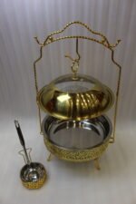 Luxury gold and stainless steel round chafing dish buffet server with a glass dome lid suspended on a gold frame, matching ladle, and buffet burner on a kitchen counter.